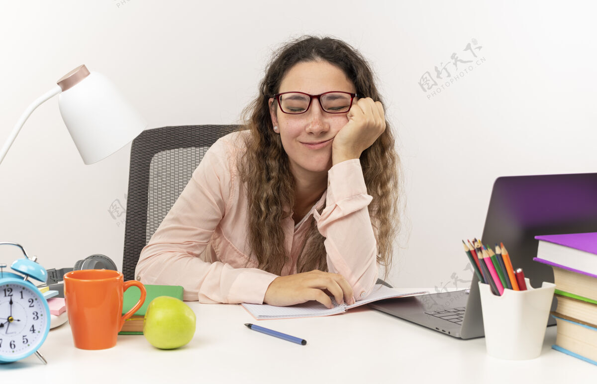 桌子高兴的年轻漂亮的女学生戴着眼镜坐在书桌旁 手里拿着学习工具做作业 手放在脸颊上 闭着眼睛孤立地站在白色的墙上人坐着眼睛