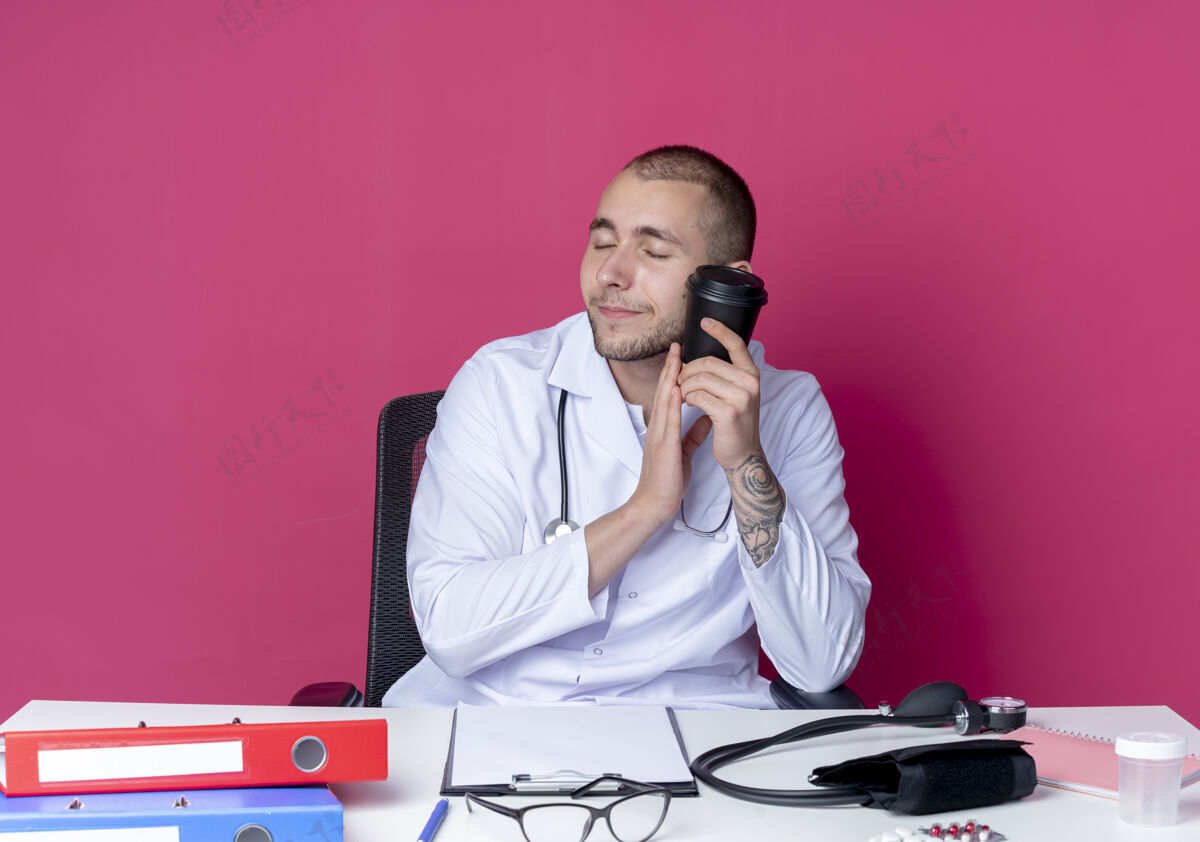 穿年轻的男医生穿着医用长袍 手持听诊器坐在办公桌旁 手里拿着工作工具 拿着塑料咖啡杯 闭着眼睛抚摸着脸 隔离在粉红色的墙上触摸抱着封闭