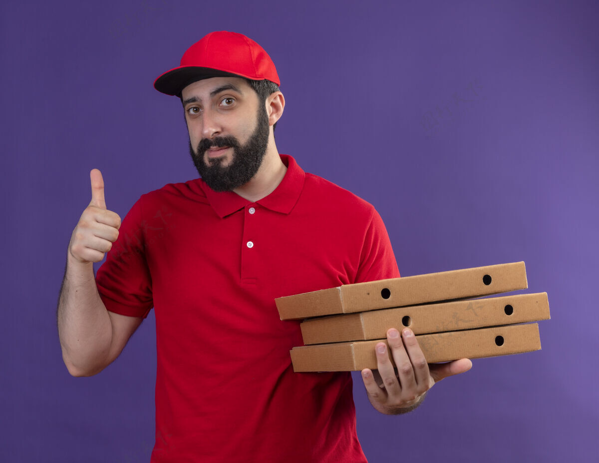 盒子年轻帅气的送货员穿着红色制服 戴着帽子 手里拿着披萨盒 在紫色的墙上孤立地竖起大拇指衣服人表情