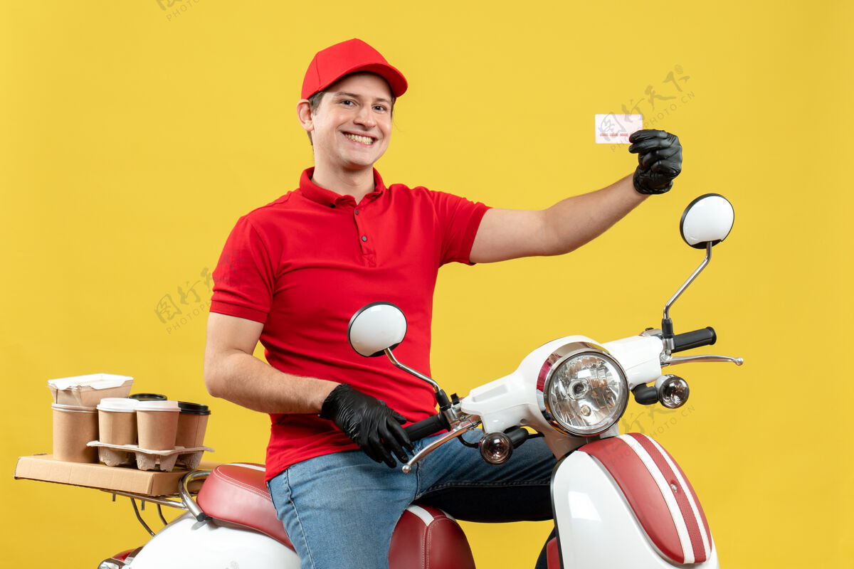 人前视图的快递员身穿红色上衣 戴着帽子手套 戴着医用口罩 坐在摩托车上拿着银行卡递送订单手套车辆银行