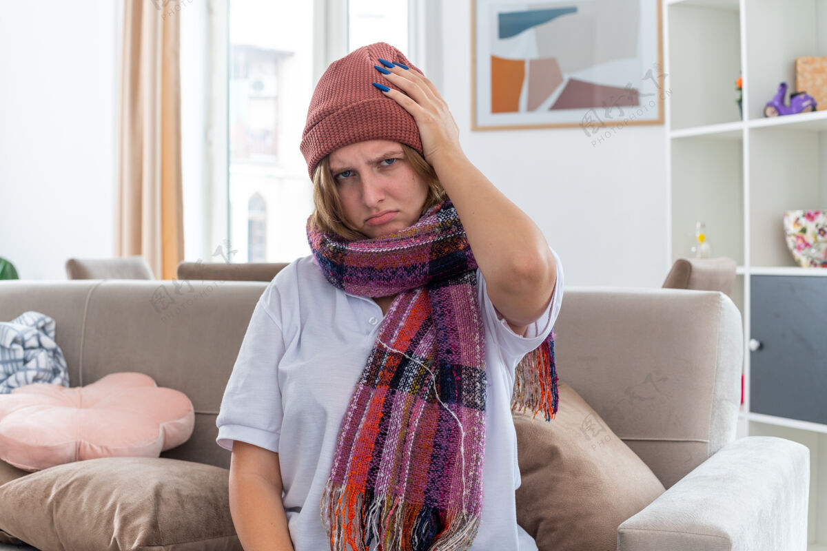 暖不健康的年轻女子戴着暖和的帽子 脖子上围着围巾 感觉不舒服 生病 患感冒和流感 手碰到头 头痛 坐在客厅的沙发上头坐周围