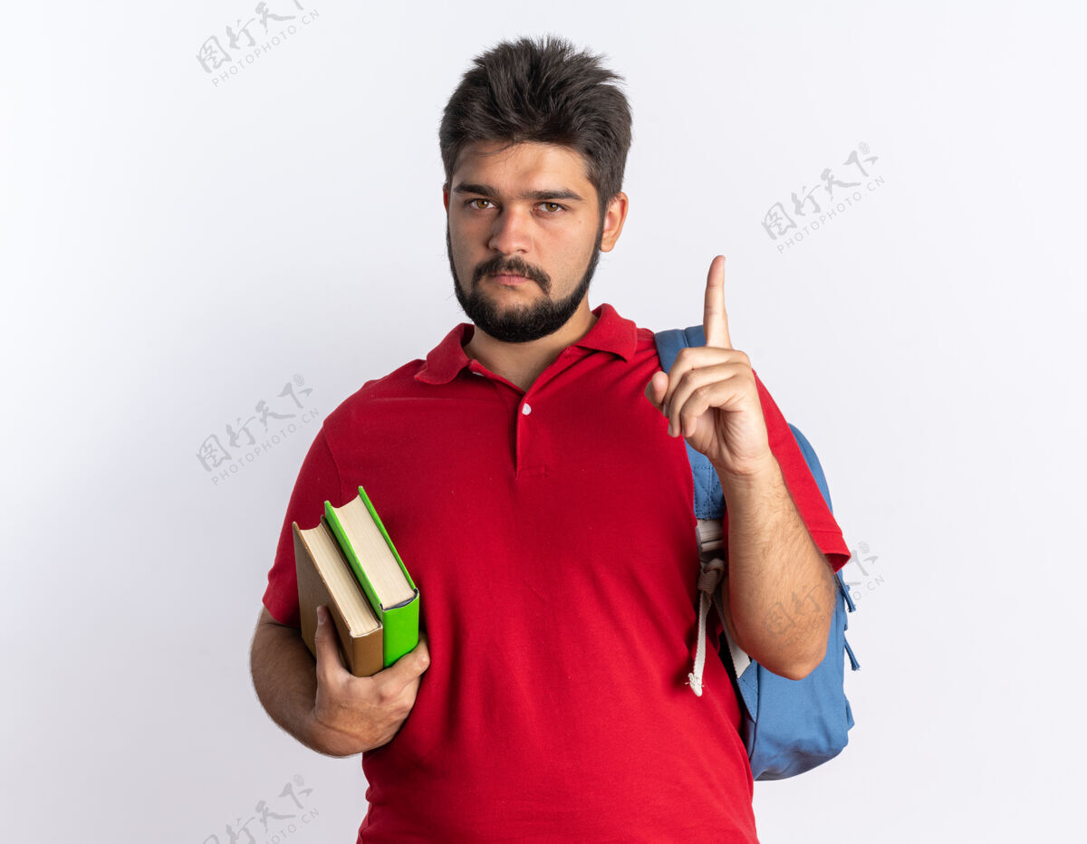 背包一个留着胡子的年轻学生 穿着红色马球衫 背着背包 拿着笔记本 严肃地看着镜头 食指站在白色背景上学生手指马球