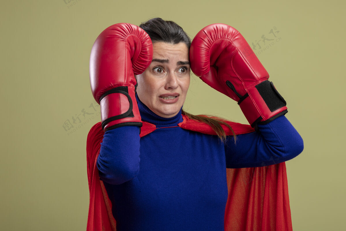 拳击害怕的年轻超级英雄女孩一边看一边戴着拳击手套把手放在头上隔离在橄榄绿上手穿年轻