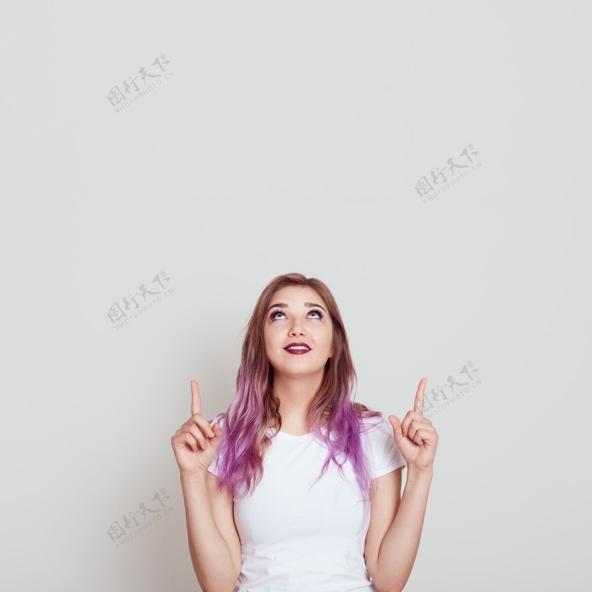 快乐穿着白色休闲t恤的年轻迷人的女士 用食指向上看 为广告或促销文本复制空间 在灰色的墙上摆出孤立的姿势年轻情绪人