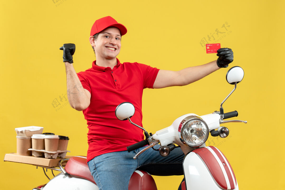 红色正面图：微笑的快递员身穿红色上衣 戴着帽子手套 戴着医用面罩 坐在滑板车上 指着后面的银行卡面具指向车辆