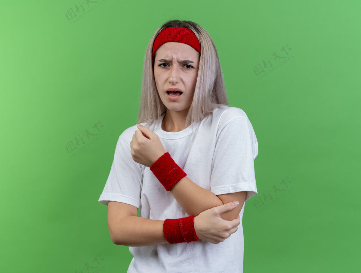 戴带着护腕戴着头带和腕带的疼痛的年轻运动女性把肘部隔离在绿色的墙上头带年轻人女孩