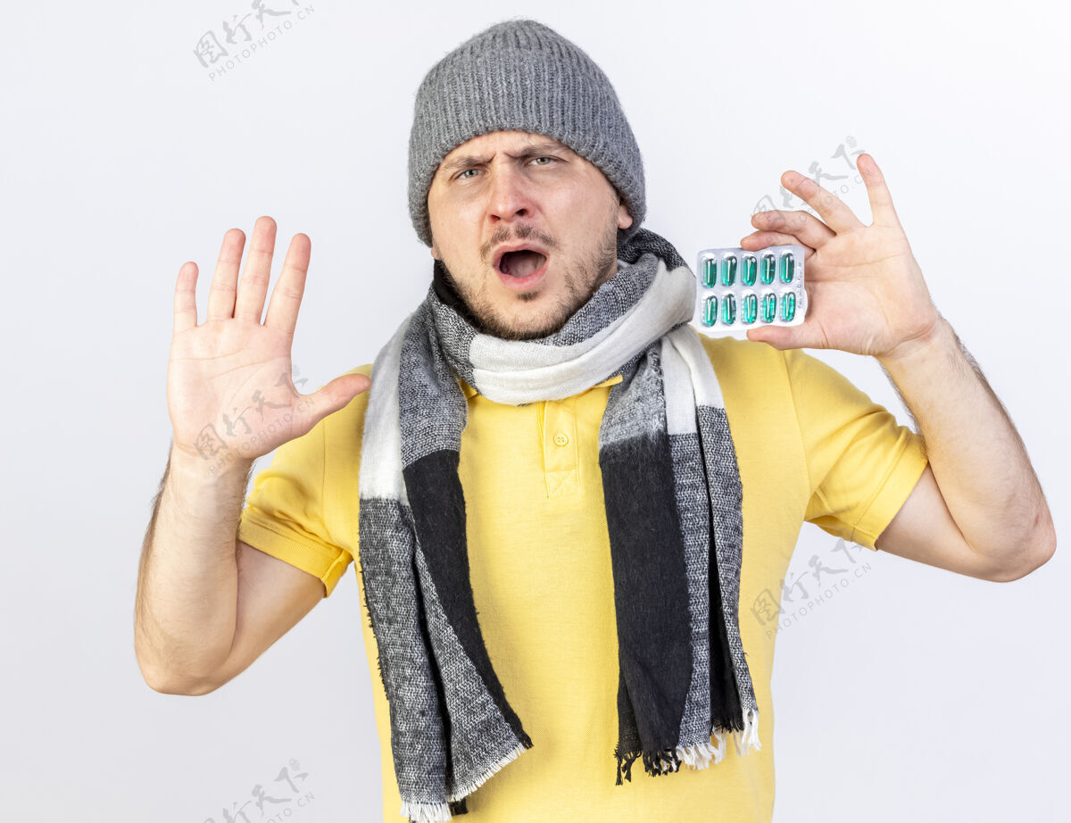 抱着年轻的金发病斯拉夫男子戴着冬天的帽子和围巾站起来 举起手来 拿着一包药片隔离在白色的墙壁与复制空间不愉快戴着帽子