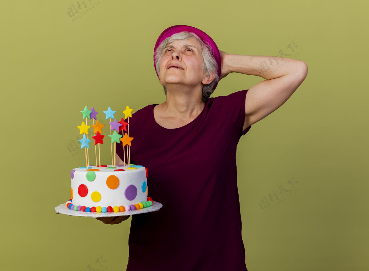 女人恼怒的戴着派对帽的老妇人把手放在头上 身后拿着生日蛋糕 隔离在橄榄绿的墙上 留着复制空间头老人橄榄