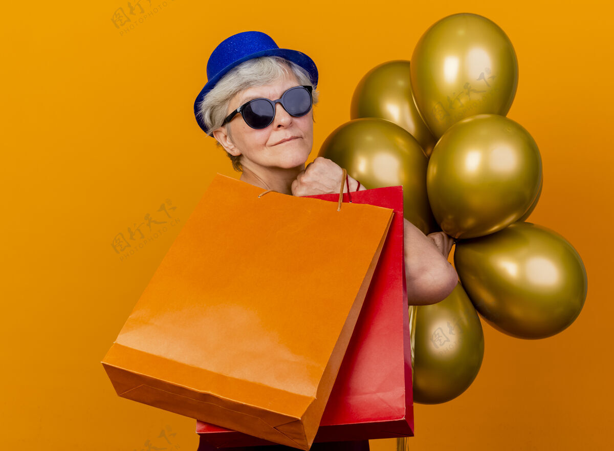 穿戴着派对帽 戴着太阳眼镜的高兴的老妇人站在一旁 手里拿着氦气球和纸购物袋 隔离在橙色的墙上 还有复印空间帽子气球老人