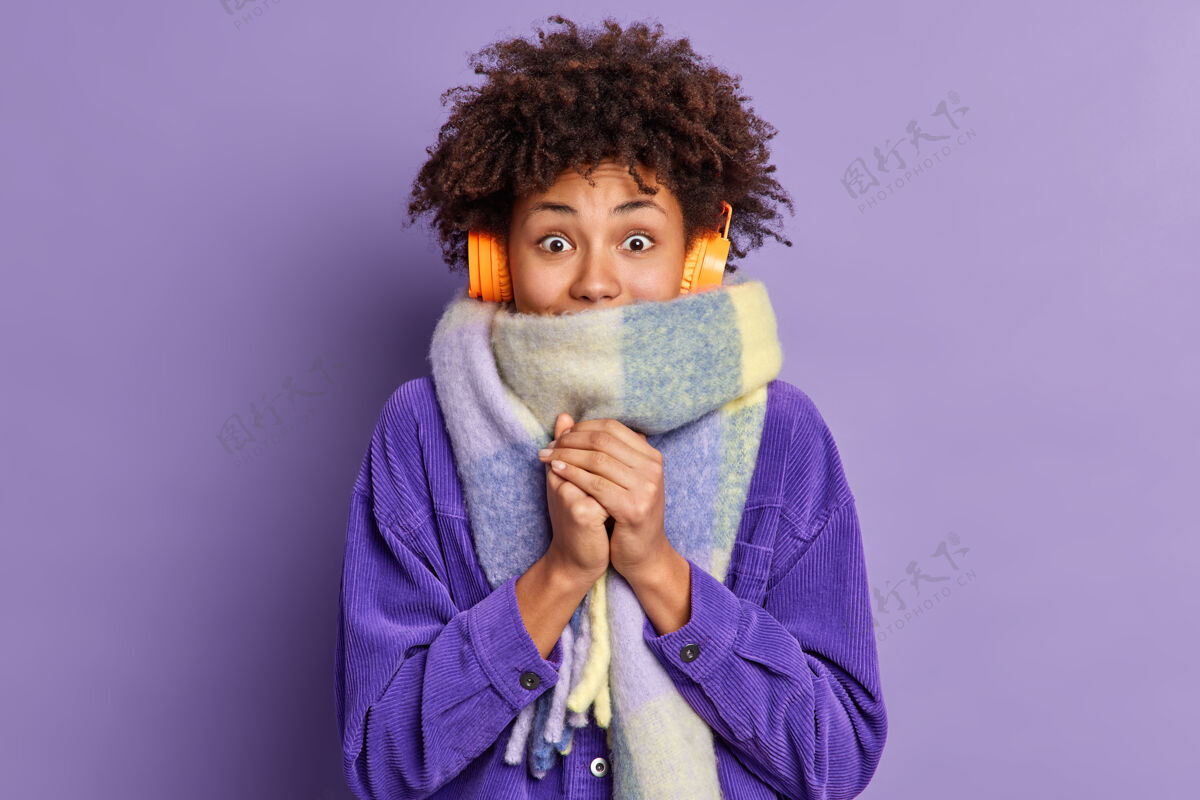 美丽美国黑人妇女在寒冷的天气里感觉很冷 冬天在街上穿着紫色夹克 围着暖和的围巾 通过无线耳机听音乐 双手合十自然非洲年轻