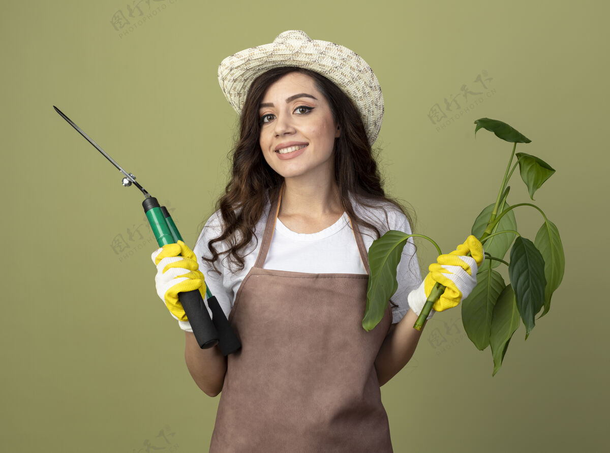 帽子微笑着的年轻女园丁穿着制服 戴着园艺帽和手套 拿着植物和园艺剪 隔离在橄榄绿的墙上 留着复制空间持有剪子年轻