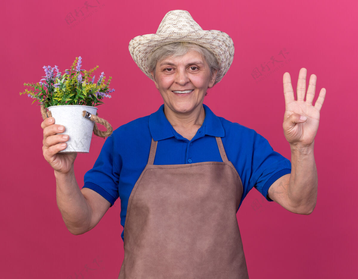 四微笑的上了年纪的女园丁戴着园艺帽 手里拿着花盆 四个手指在粉色上做手势四四四