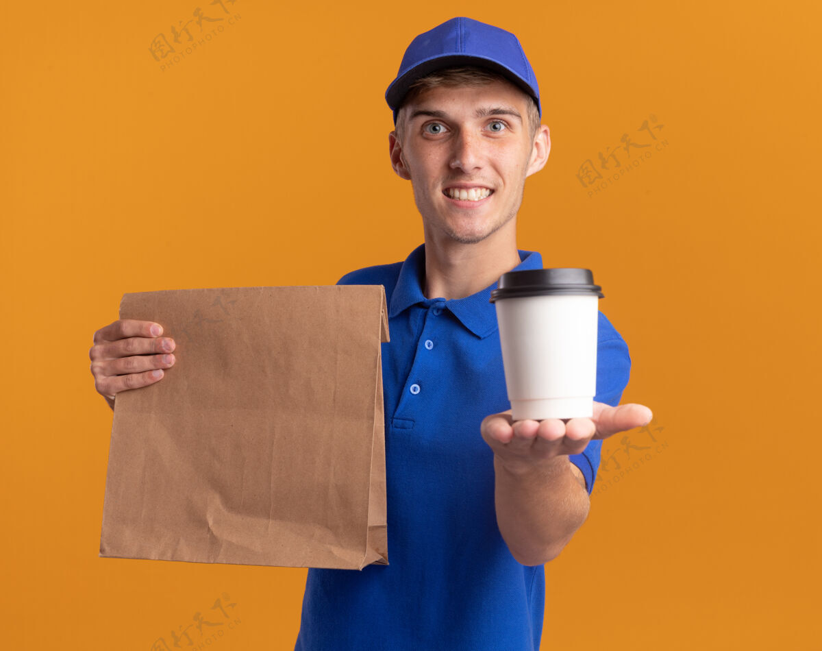 年轻微笑着的年轻金发送货男孩拿着纸包装和杯子在橙色送货金发纸