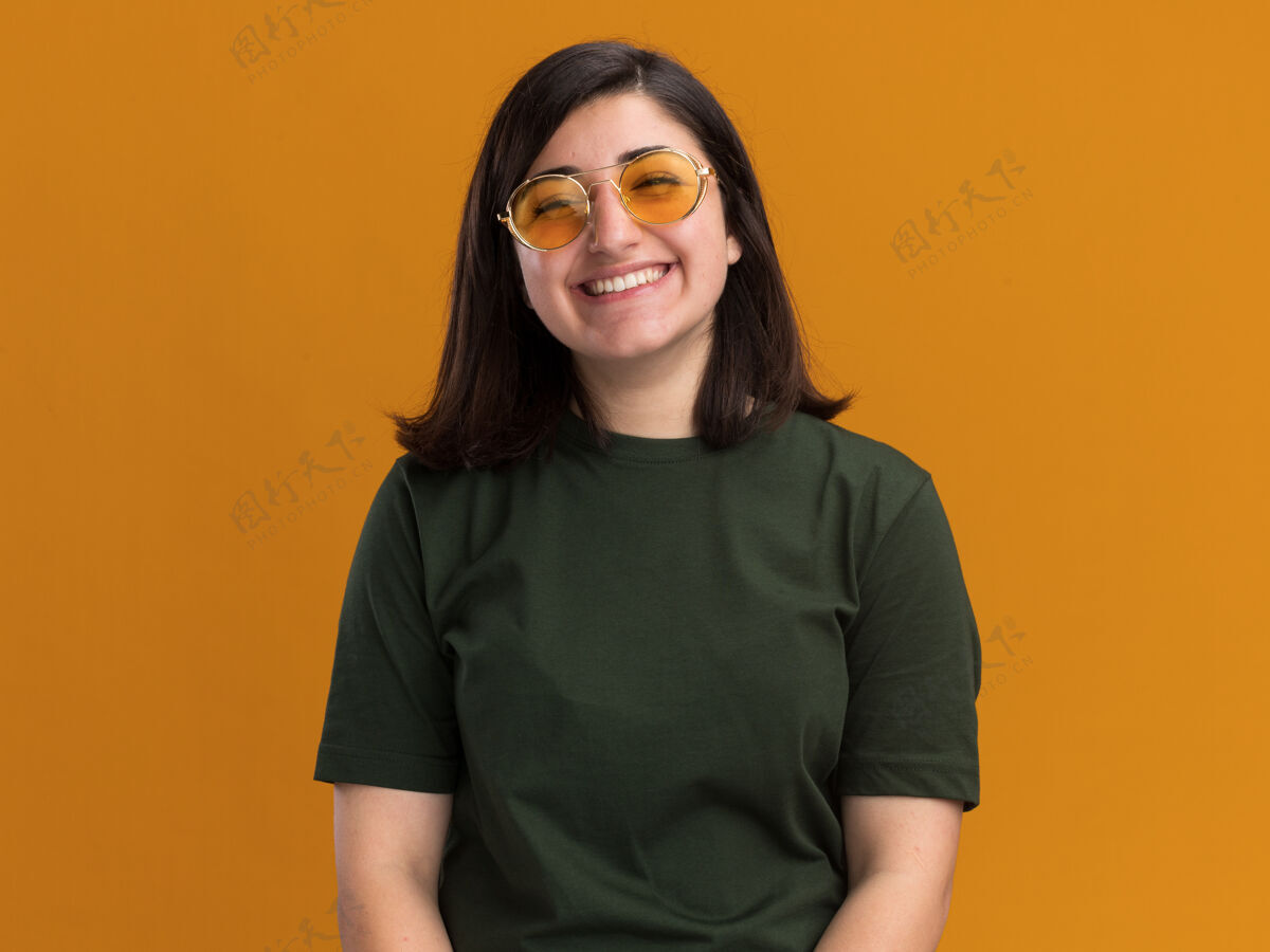 微笑微笑着的年轻漂亮的高加索女孩戴着太阳眼镜看着橙色的相机橙色相机年轻