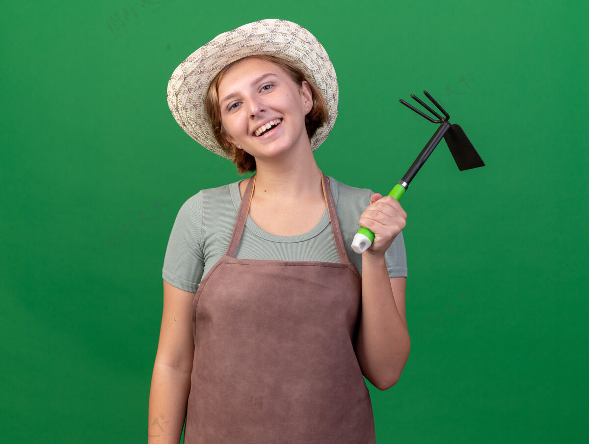 女的微笑的年轻斯拉夫女园丁戴着园艺帽拿着锄头耙在草地上斯拉夫人帽子花园