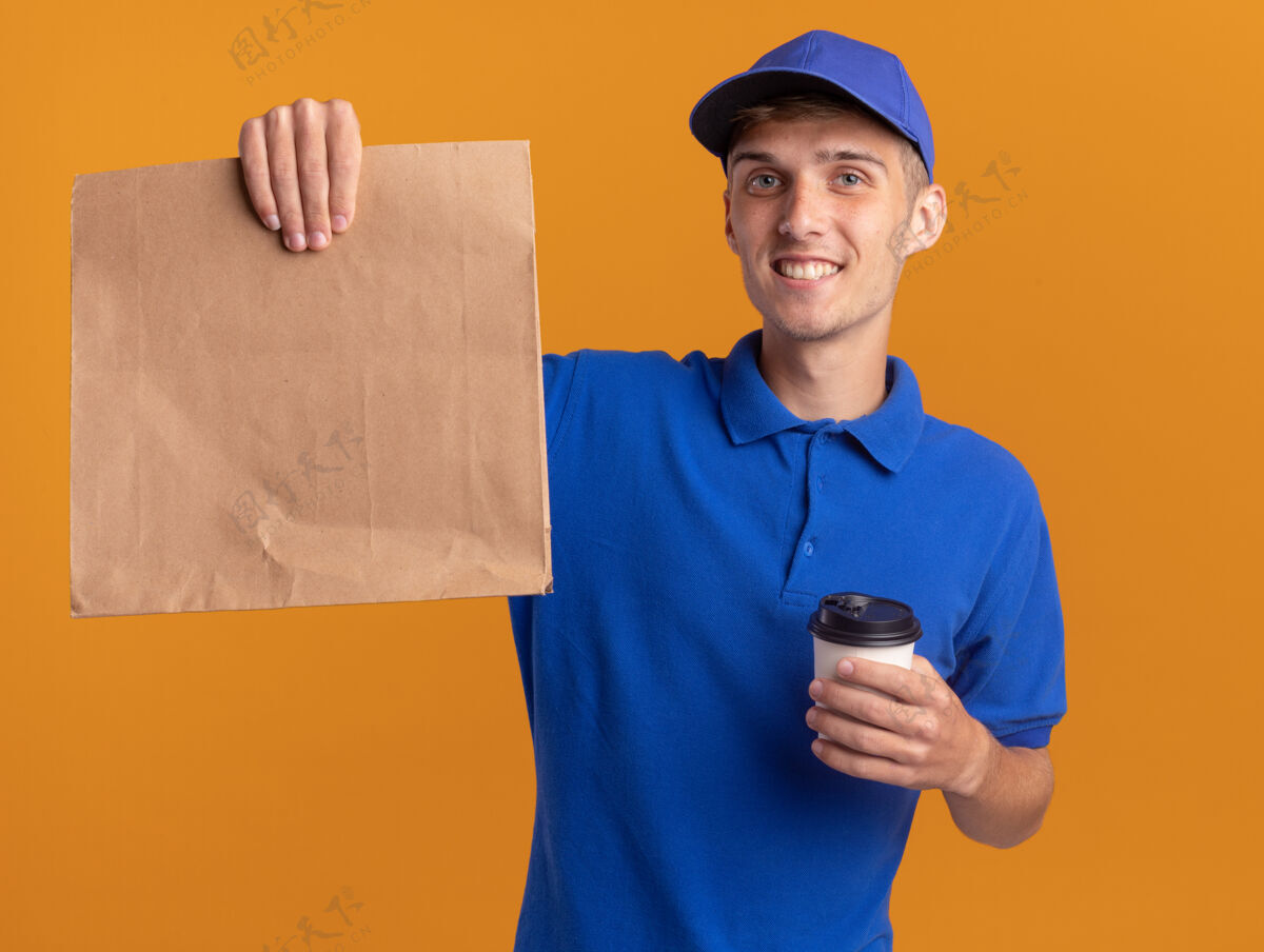 微笑微笑着的年轻金发送货男孩拿着纸包装和外卖杯在橙色送货年轻金发