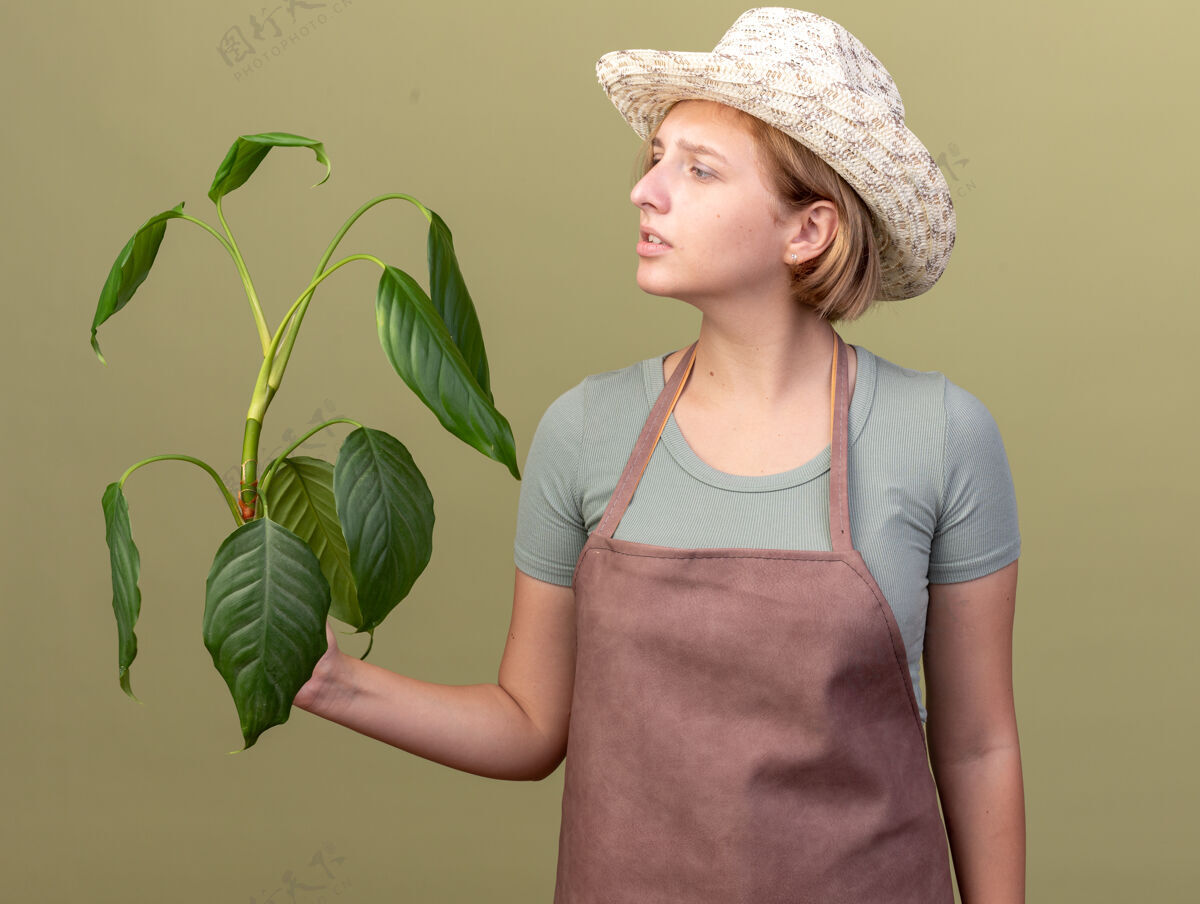 帽子不知所措的年轻斯拉夫女园丁戴着园艺帽拿着橄榄绿上的植物看着无知园艺女性