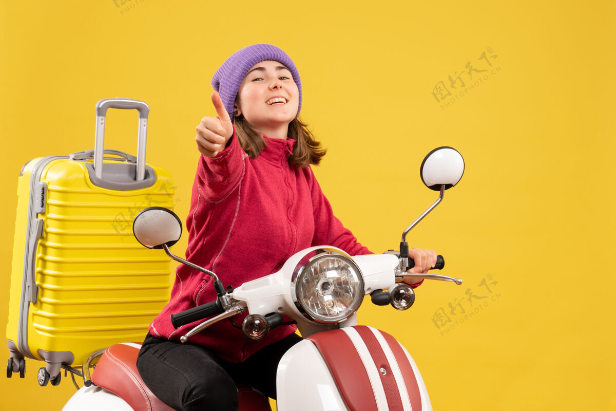 肖像正面图骑着轻便摩托车的快乐的年轻女子竖起大拇指前面人轻便摩托车