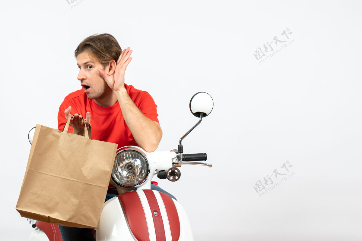 八卦穿着红色制服的年轻送货员坐在踏板车上 手里拿着纸袋 听着白墙上的最后一句闲话送货员人滑板车