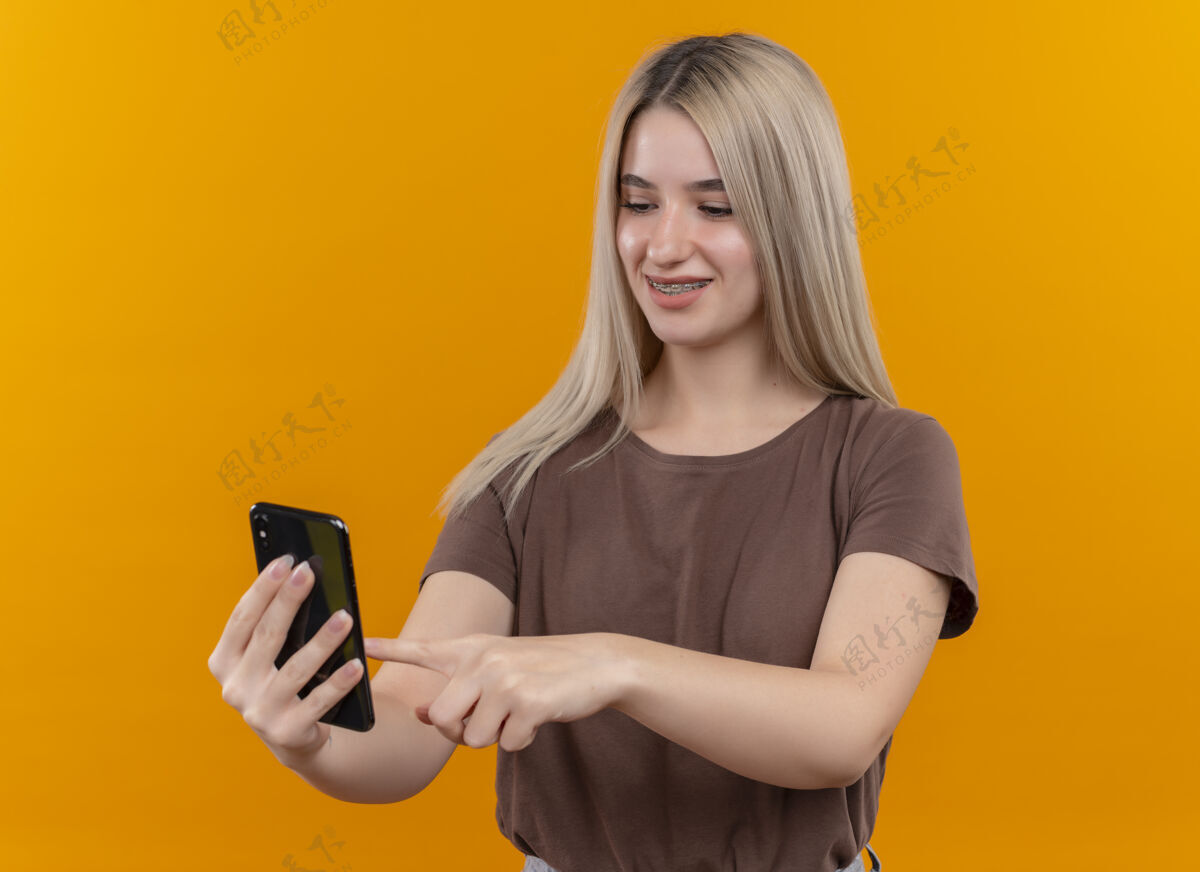 微笑微笑着戴着牙套的金发女孩拿着手机 用手指触摸着孤立的橙色空间电话女孩手指