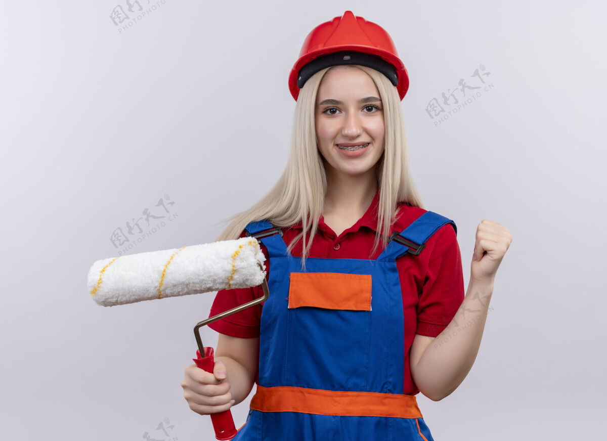 女孩微笑着的年轻金发工程师建筑工女孩穿着制服 戴着牙套 拿着油漆辊 举起拳头对着孤立的白色空间金发微笑工程师