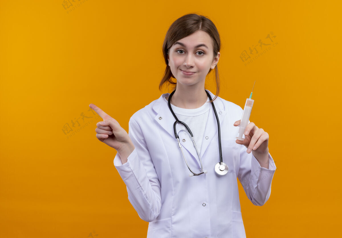 医疗高兴的年轻女医生穿着医用长袍和听诊器拿着注射器 指着左边孤立的橙色空间和复印空间手持年轻长袍