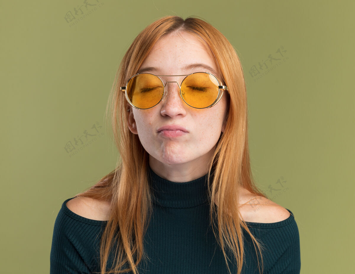 雀斑失望的年轻红发姜女 戴着太阳眼镜 满脸雀斑 闭着眼睛站在橄榄绿上眼睛眼镜失望