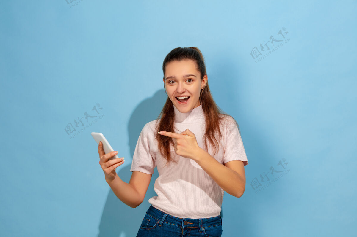 面部指着智能手机 微笑着蓝色背景上的白人少女肖像穿着休闲服的漂亮模特人类情感的概念 面部表情 销售 广告看起来很开心学习手财务