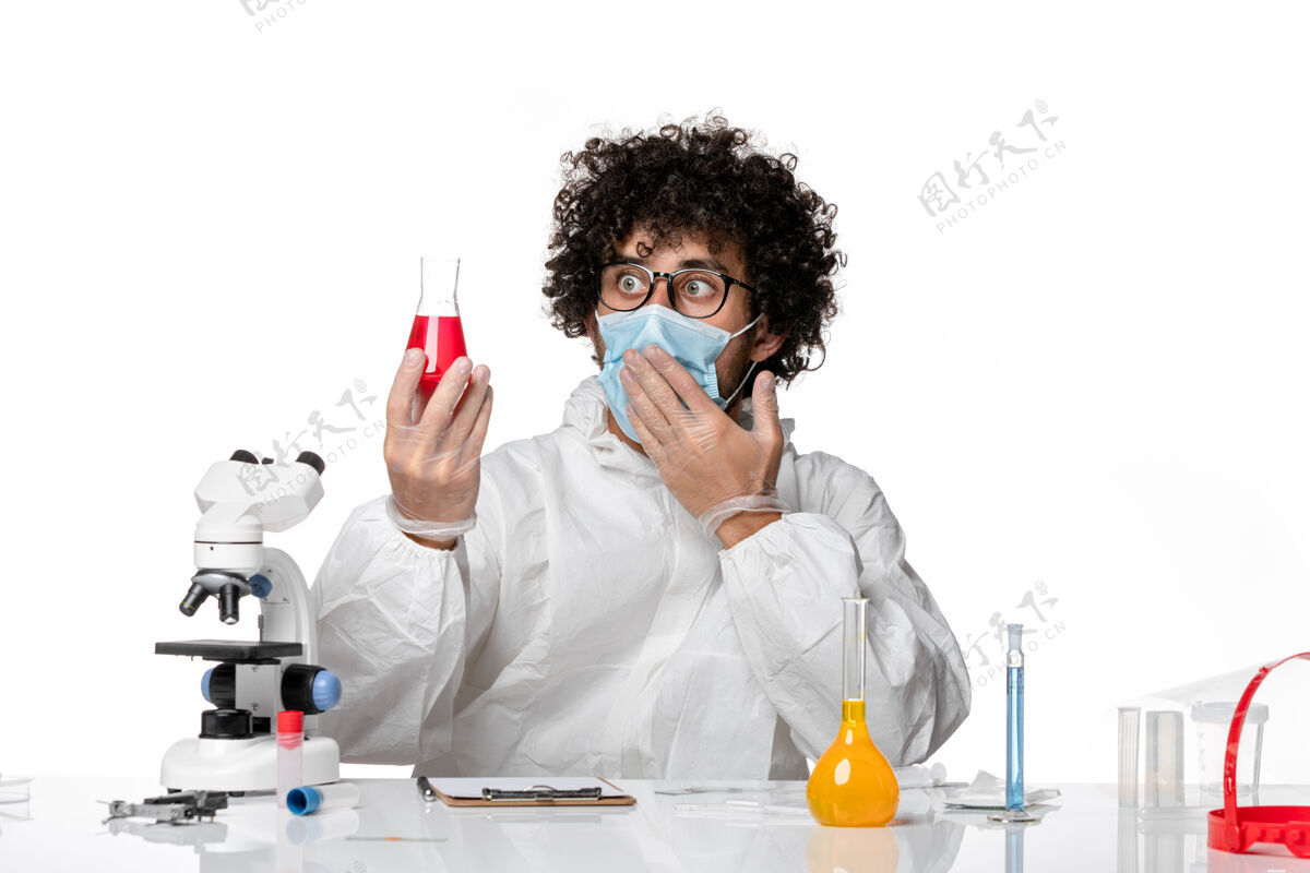 口罩正面图：男医生穿着防护服 戴着口罩 白色桌子上拿着装有红色溶液的烧瓶大流行视图外套