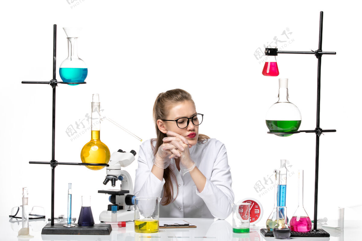 专业前视图穿着白色医疗服的女医生坐在桌子前 拿着白色背景的病毒化学大流行病毒的解决方案医生套装医学