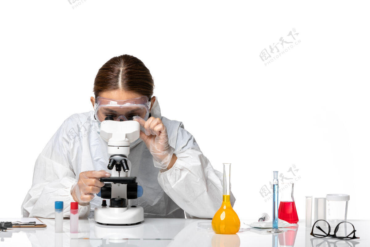 药品前视女医生穿着特制的西服 戴着口罩 用显微镜对着白色办公桌上的冠状病毒大流行女医生面罩人