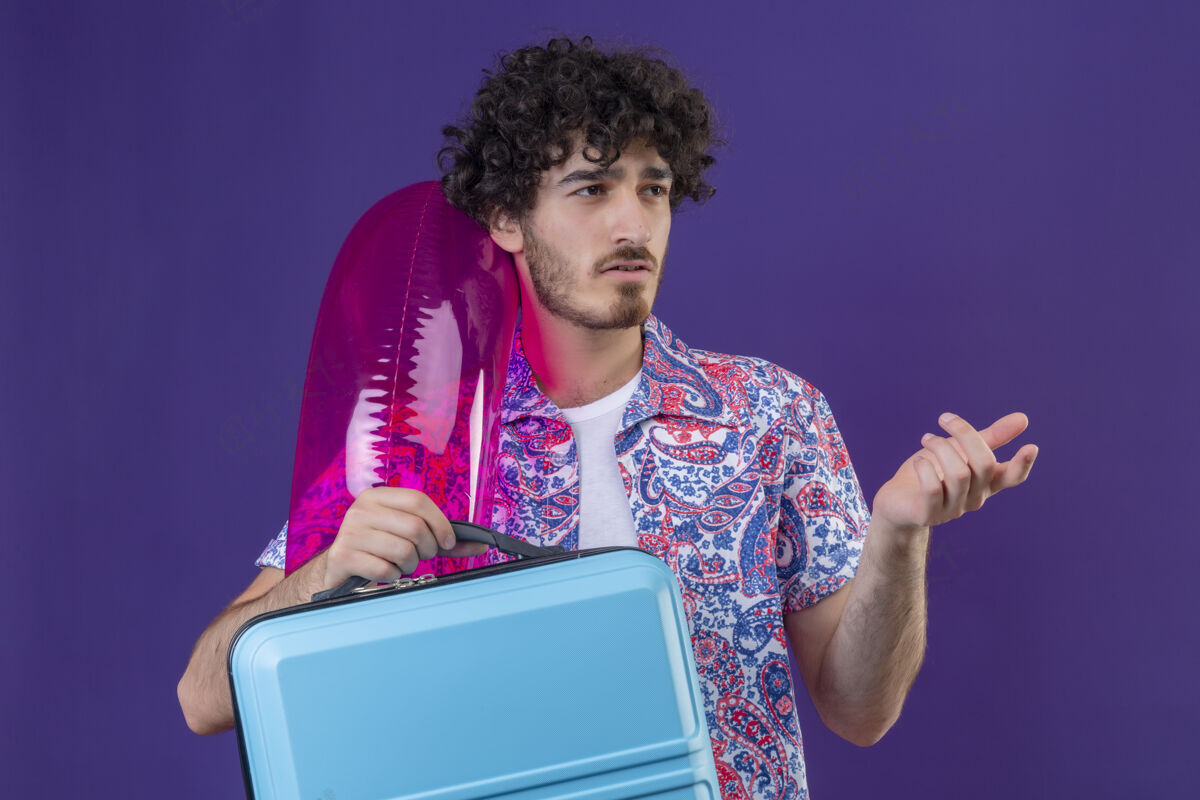 年轻困惑的年轻英俊的卷发旅行者男子拿着手提箱和游泳圈指着右边孤立的紫色空间手提箱侧身抱着