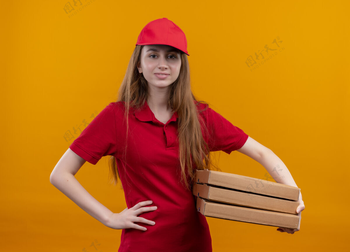 橙色在孤立的橙色空间里 穿着红色制服的自信的年轻送货女孩手放在腰上拿着包裹腰手信心