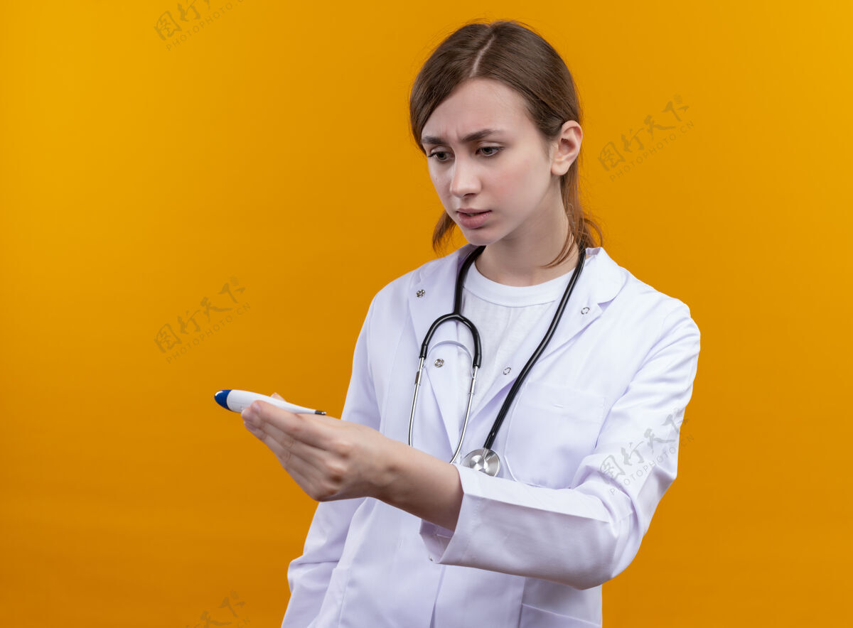 焦虑焦虑的年轻女医生穿着医用长袍 手持听诊器 拿着体温计 在孤立的橙色空间里看着它长袍体温计医疗