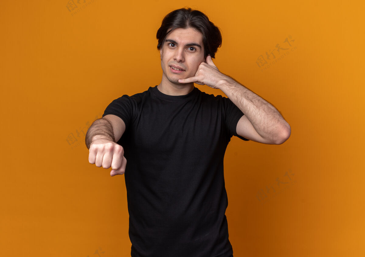 人一个穿着黑色t恤的年轻帅哥 在橙色的墙上展示着打电话的姿势 并在前面伸出拳头抱着手势姿势
