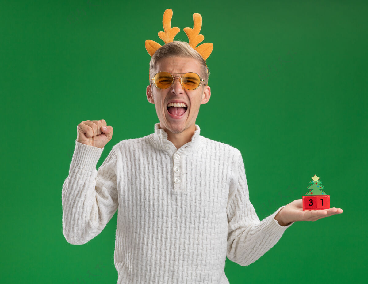 圣诞快乐快乐的年轻帅哥戴着驯鹿鹿角头带戴着眼镜拿着圣诞树玩具和日期看着相机做“是”的手势隔离在绿色背景上绿色举行家伙