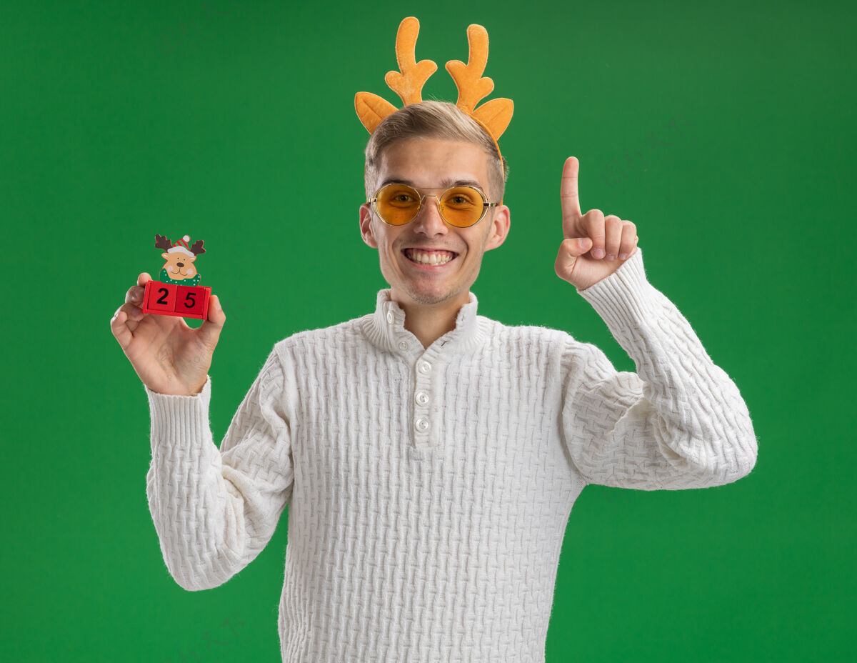 眼镜快乐的年轻帅哥戴着驯鹿鹿角头带戴着眼镜拿着圣诞树玩具和日期看着相机指着绿色背景上孤立的家伙日期圣诞快乐