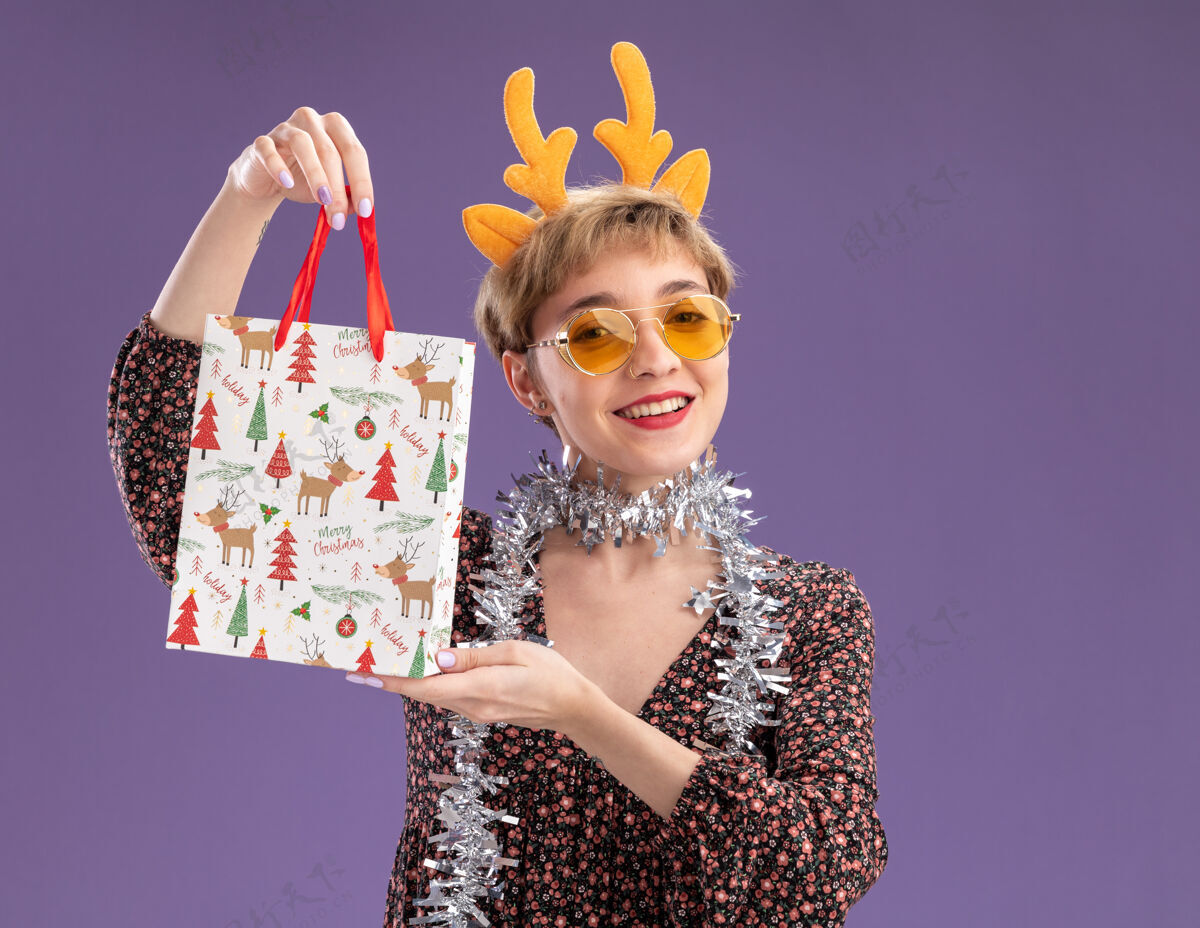 花环快乐的年轻漂亮女孩戴着驯鹿鹿角头带 脖子上戴着金箔花环 戴着眼镜 手里拿着圣诞礼品袋 看着紫色背景上孤立的相机金属丝空间头带