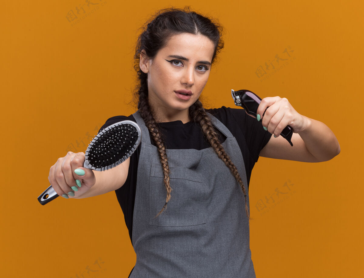 表情自信的年轻女理发师穿着制服站在战斗的姿势拿着梳子与剪子孤立在橙色墙上女性年轻人人