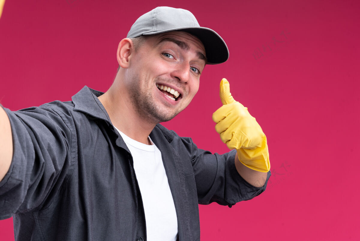 年轻人微笑着的年轻帅气的清洁工 穿着t恤 戴着帽子 戴着手套 举着大拇指站在粉红色的墙上拇指微笑表情