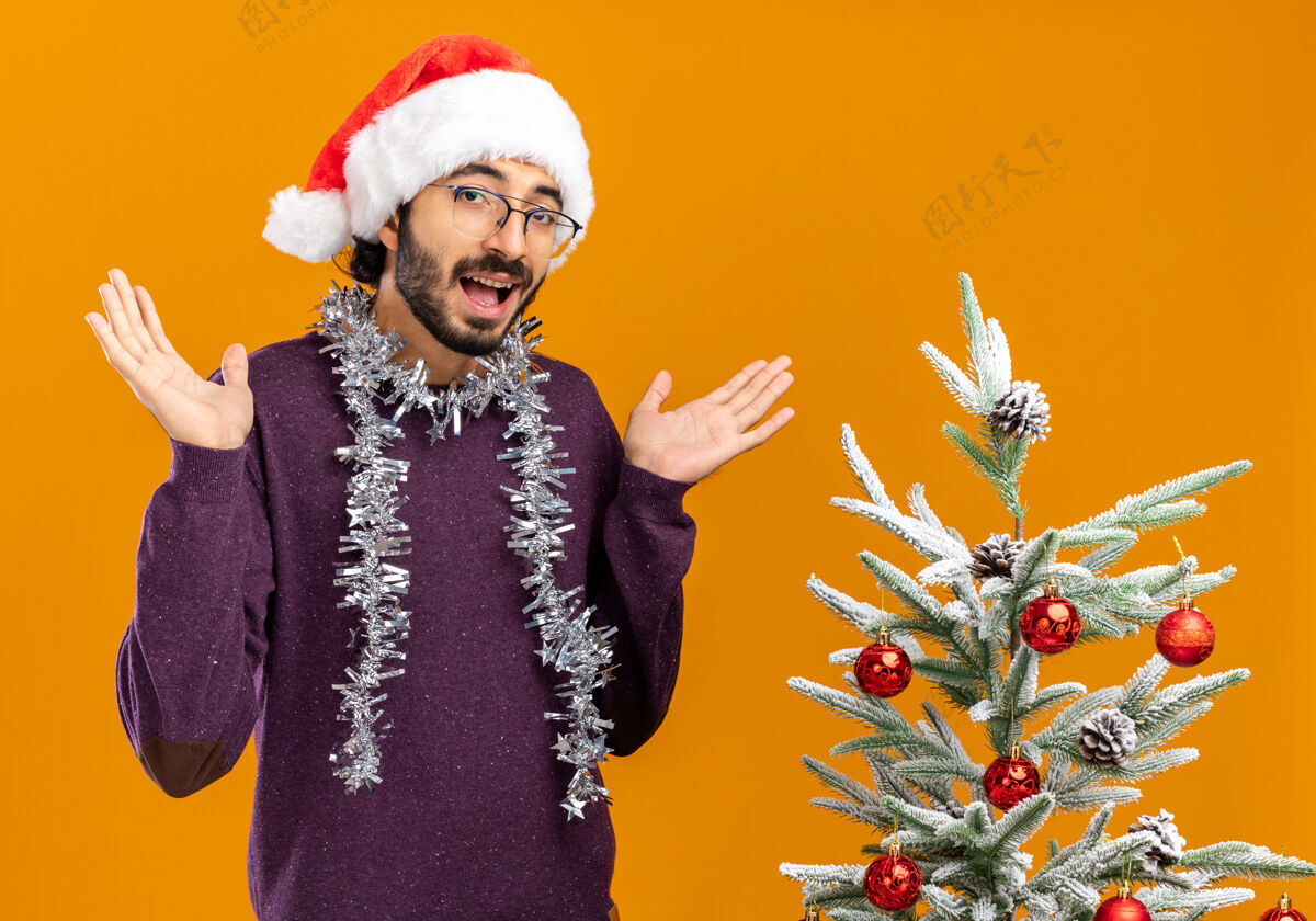 手快乐的年轻帅哥站在圣诞树旁 戴着圣诞帽 脖子上戴着花环 双手分开放在橙色的背景上圣诞快乐年轻人橘子