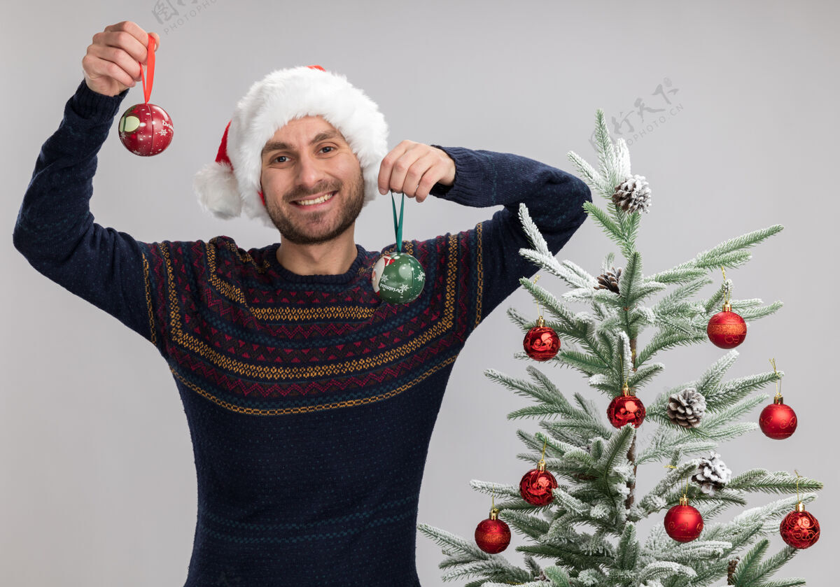 圣诞树快乐的年轻白人男子戴着圣诞帽站在圣诞树旁看着摄像机举起圣诞装饰球隔离在白色背景上装饰品男人球