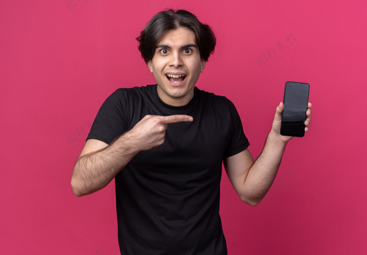 站兴奋的年轻帅哥穿着黑色t恤拿着手机指着隔离在粉红色墙上的手机市民年轻穿着