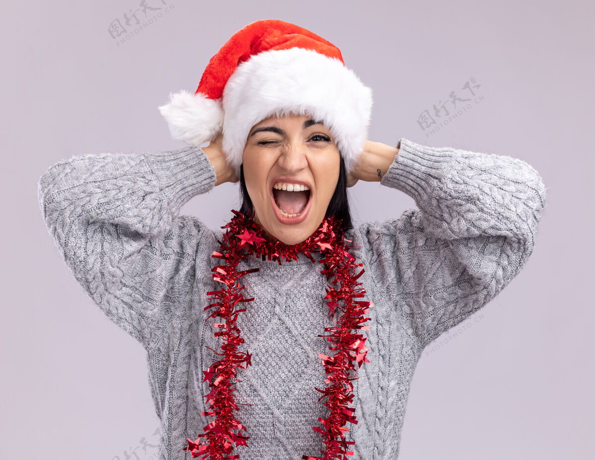 圣诞快乐快乐的年轻白人女孩戴着圣诞帽 脖子上戴着金属丝花环 看着摄像机 双手放在头上 在白色背景下尖叫和眨眼花环年轻尖叫