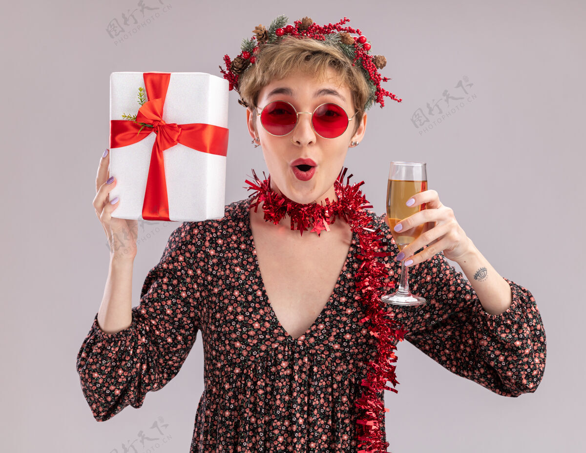 圣诞快乐给人印象深刻的年轻漂亮女孩戴着圣诞花环 脖子上戴着金箔花环 戴着眼镜拿着礼包 喝着香槟看着隔离在白色背景上的相机新年圣诞眼镜