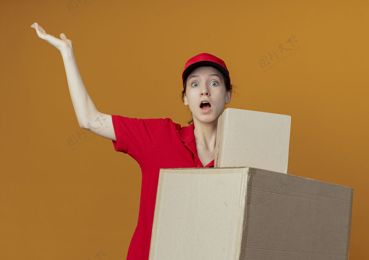漂亮令人印象深刻的年轻漂亮的送货女孩在红色制服和帽子举行纸箱和显示空的手孤立在橙色的背景盒子帽子年轻