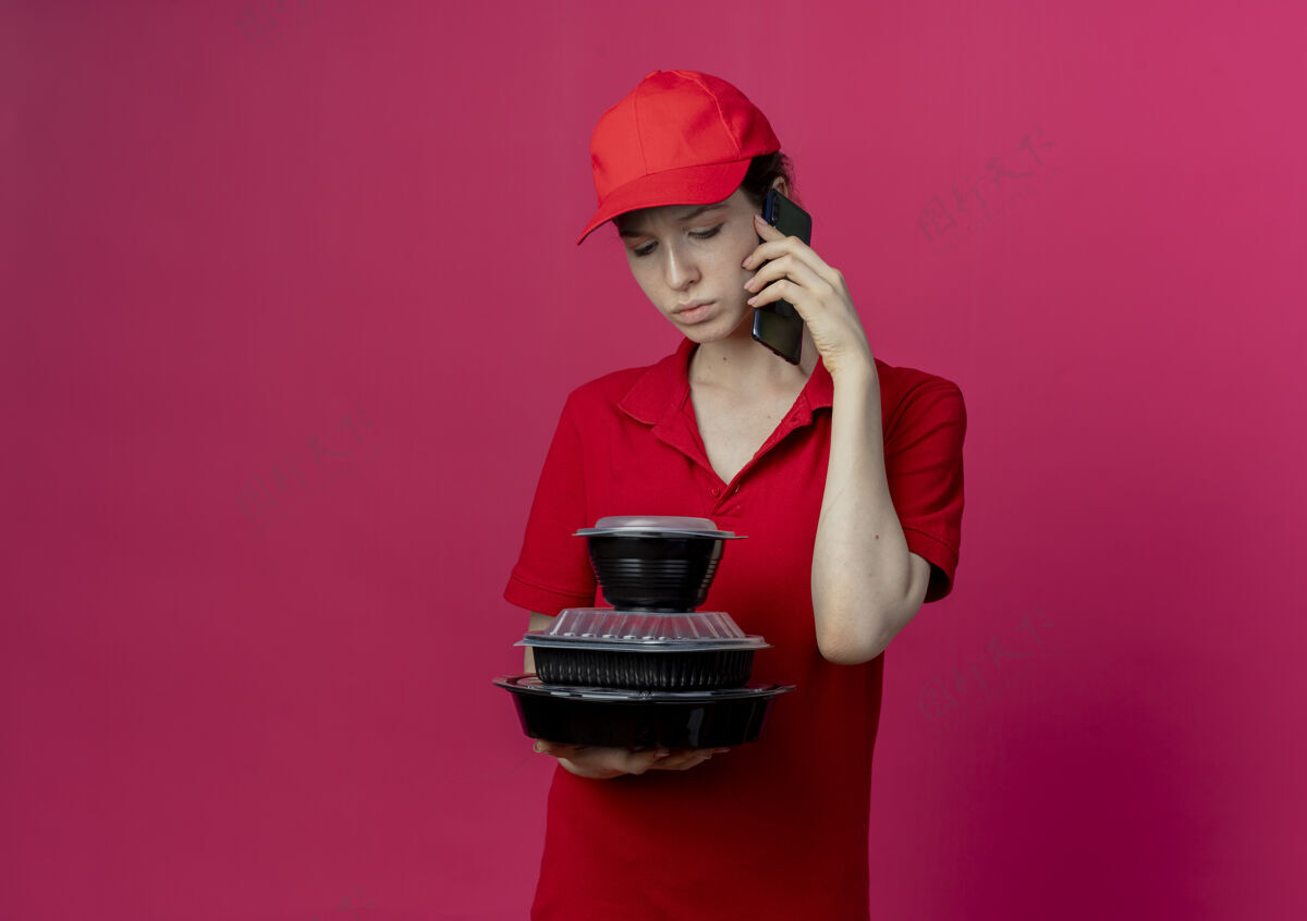 不愉快年轻漂亮的送货员 穿着红色制服 戴着帽子 一边打电话 一边拿着 一边看着被隔离在深红色背景下的食品容器 还有复印空间送货红色持有