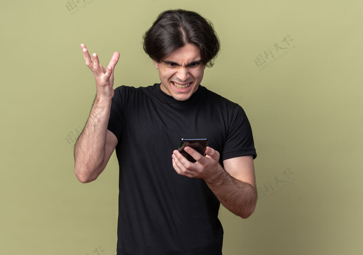 姿势愤怒的年轻帅哥身穿黑色t恤 手持手机 看着橄榄绿的墙上孤立的电话市民人人