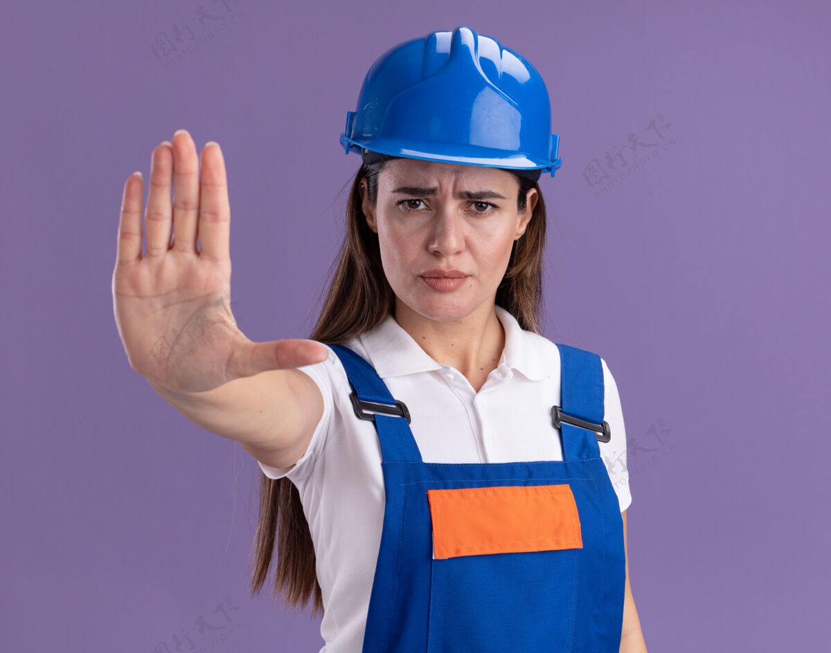 姿势穿制服的严格的年轻建设者妇女在紫色的墙上显示停止手势表情制服人