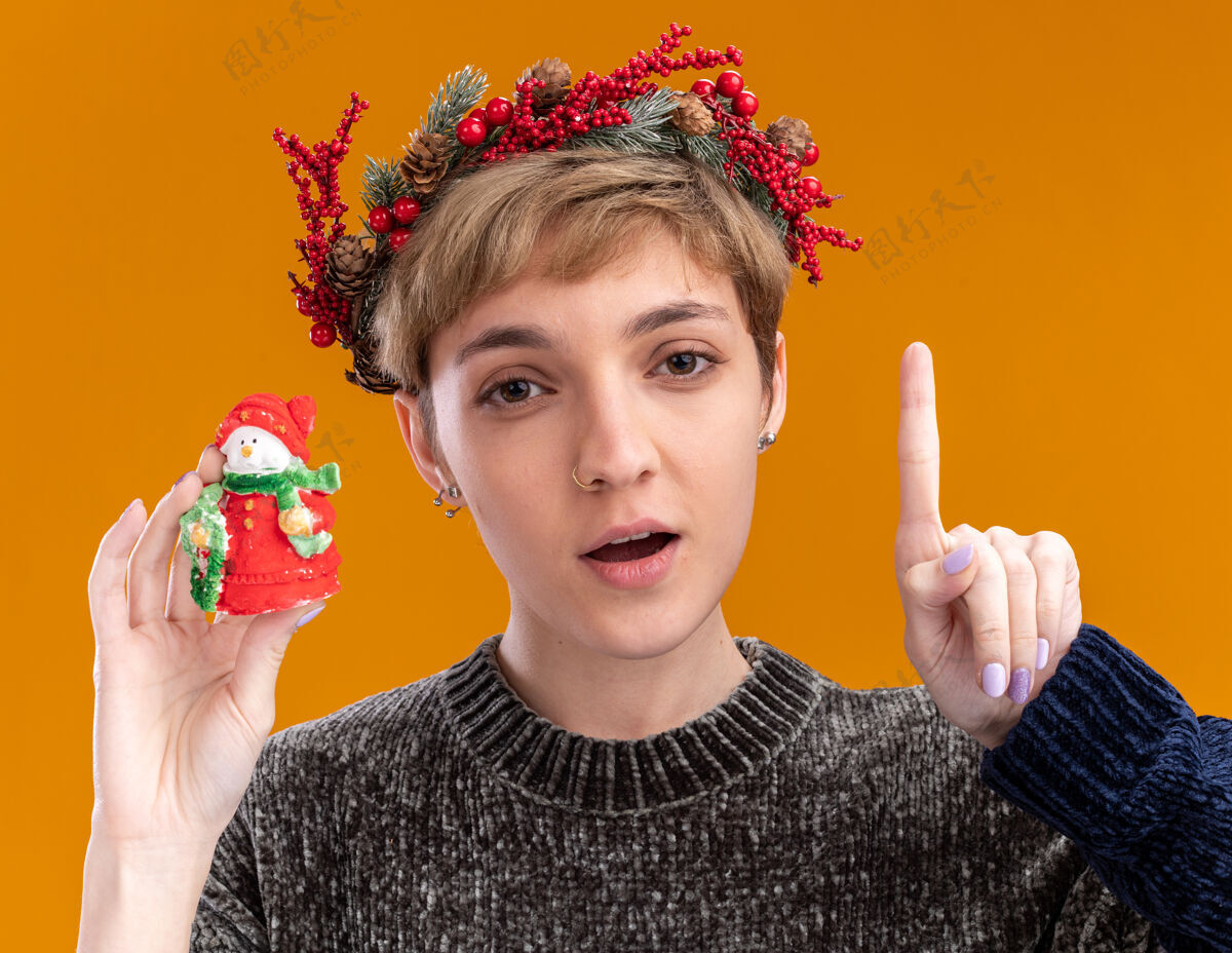 向上令人印象深刻的年轻漂亮女孩戴着圣诞花环手持小圣诞雪人雕像看着摄像机指着孤立的橙色背景圣诞节年轻雪人